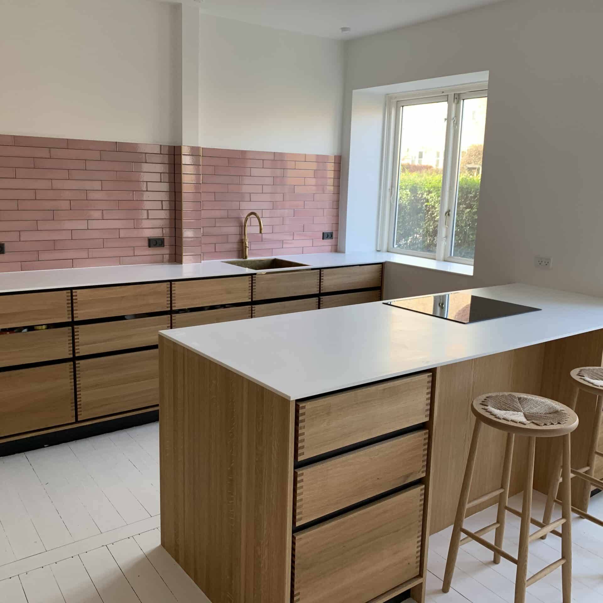Moderne køkken med lyse træskabe, hvide bordplader og pink metrofliser. Et tømrer Slagelse-touch ses i guldarmaturet og barstolene i træ ved den centrale ø. Lyst, minimalistisk design med et stort vindue.