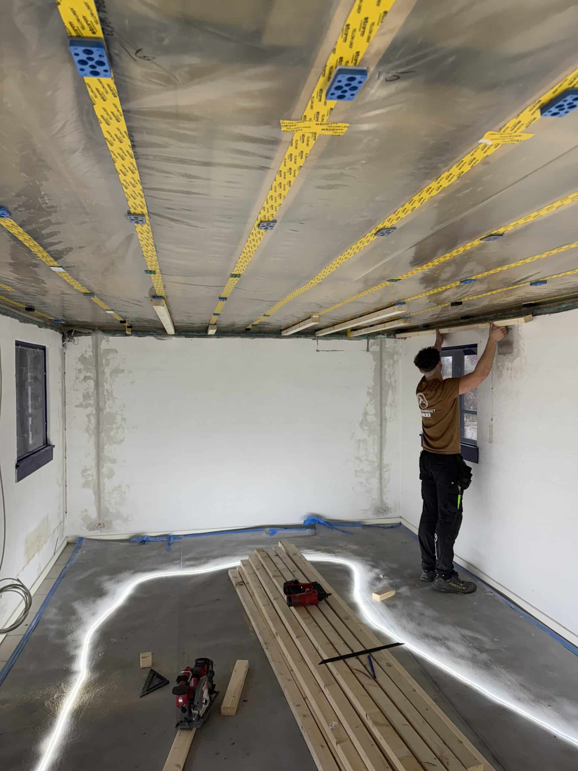 En person i Hvalsø monterer isolering og trælister på et loft i et rum under renovering, med værktøj og træplanker på gulvet og plastik, der dækker dele af loftet.