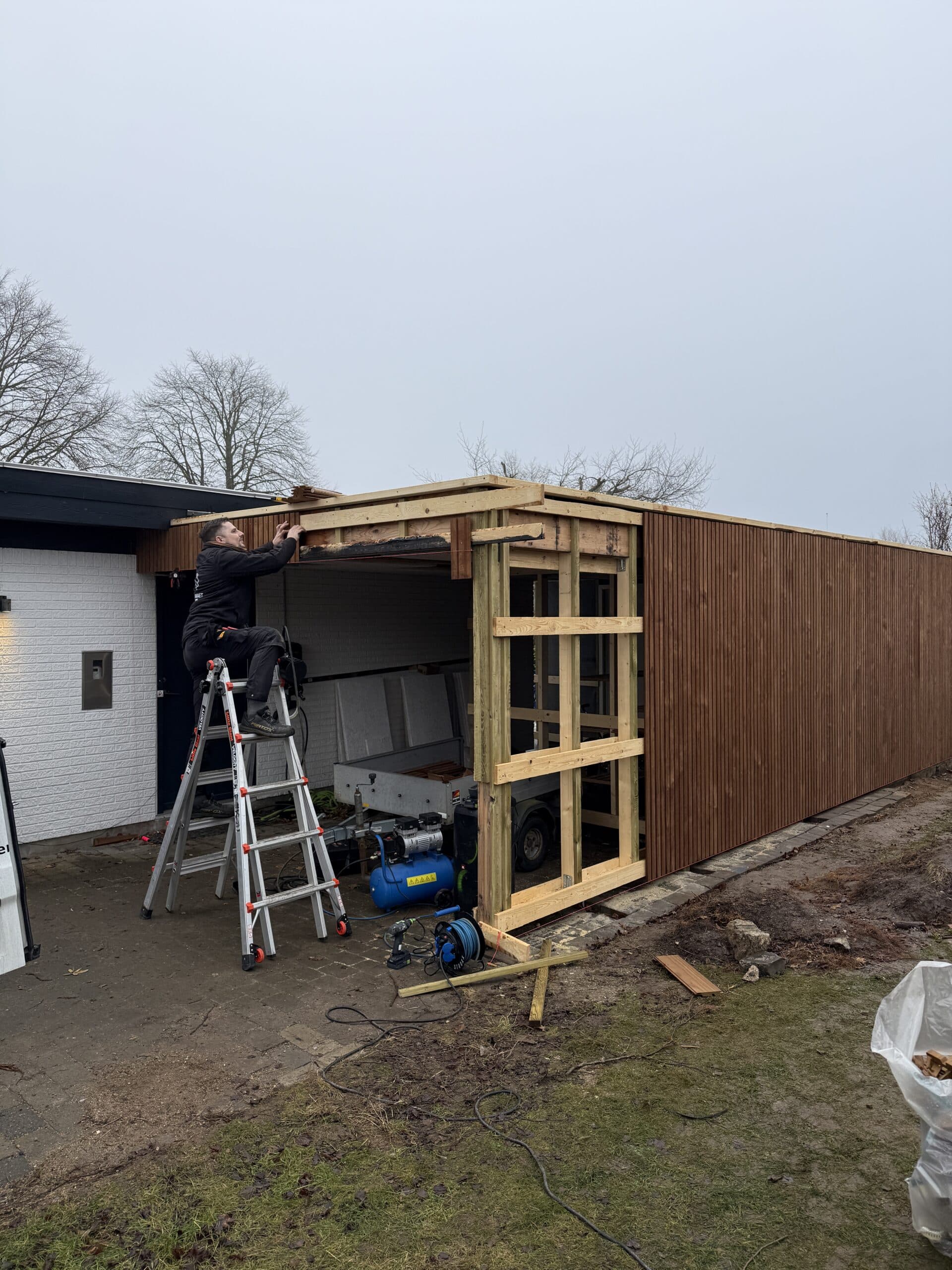 En person står på en stige i Næstved og arbejder på trærammen til en tilbygning. Byggeredskaber og materialer ligger spredt i nærheden. Strukturen har lodrette træpaneler og er knyttet til et hvidt murstenshus.