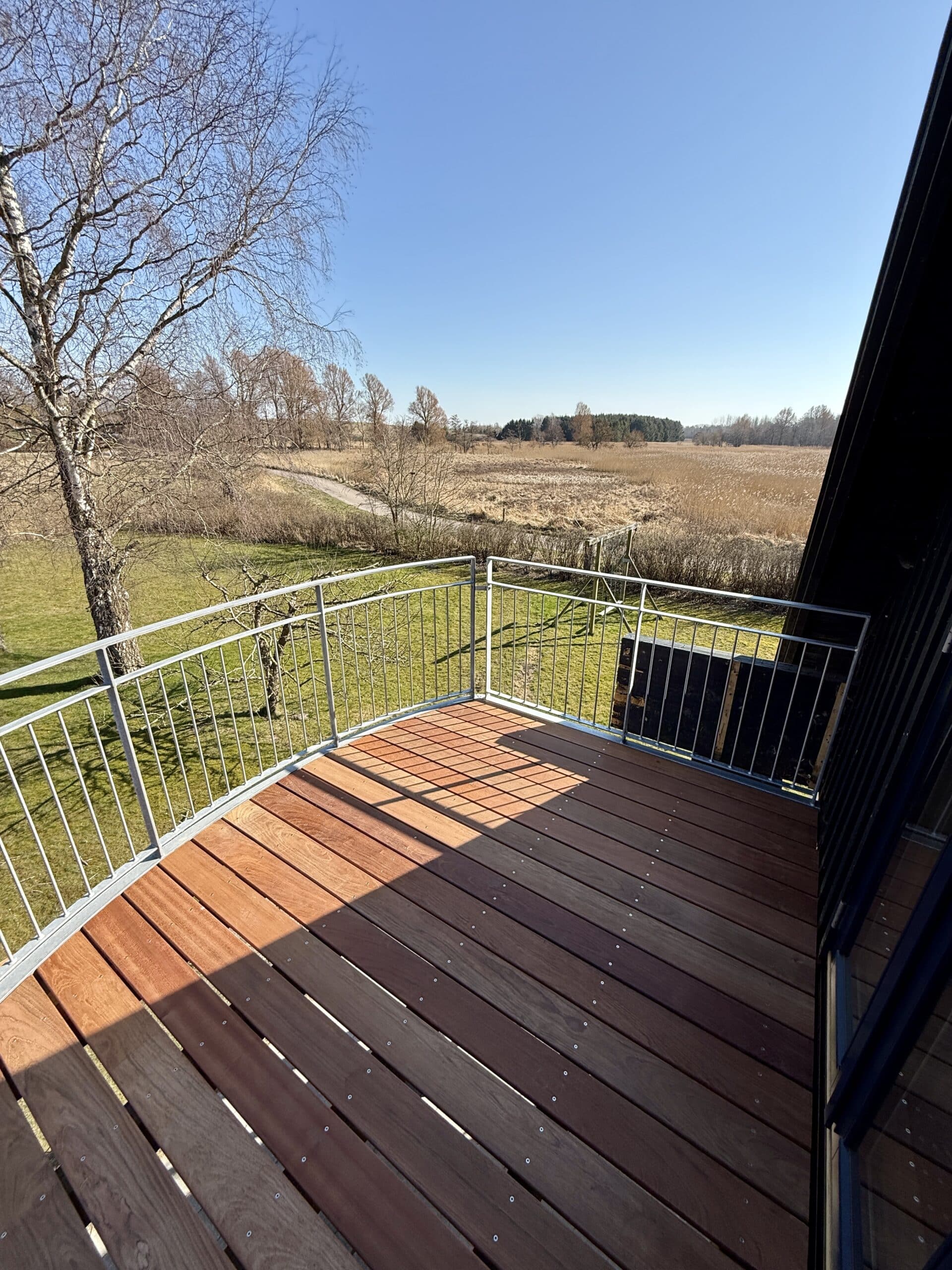 Fra en træaltan med metalrækværk er der udsigt til en græsklædt gård, nøgne træer og en åben mark under en klar blå himmel på en solskinsdag i Næstved.
