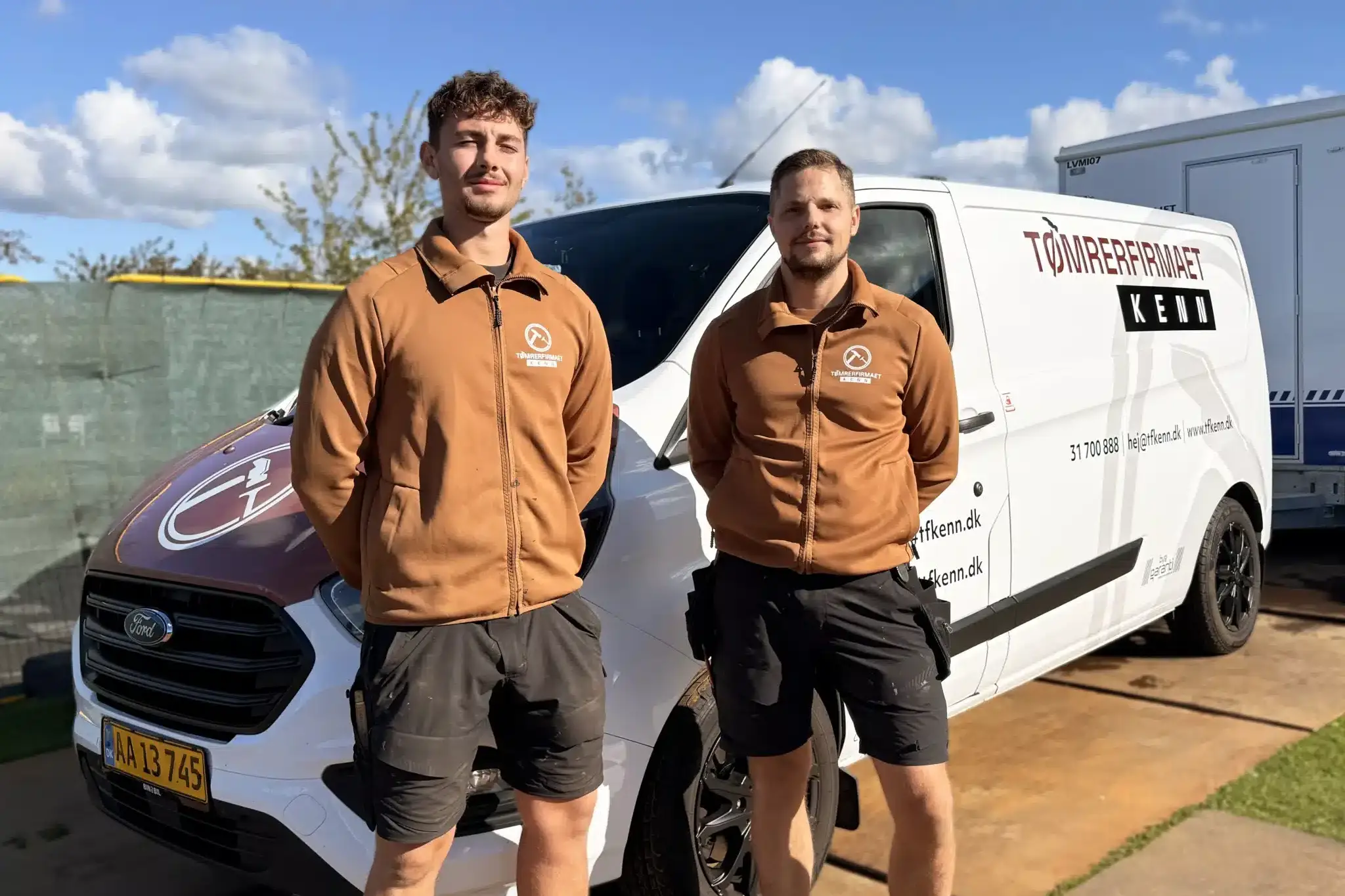 To m&aelig;nd i brune jakker og sorte shorts st&aring;r foran en hvid varevogn med T&oslash;mrerfirmaet Ken og kontaktoplysninger p&aring; siden. Scenen er udend&oslash;rs p&aring; en solrig dag med bl&aring; himmel og spredte skyer.