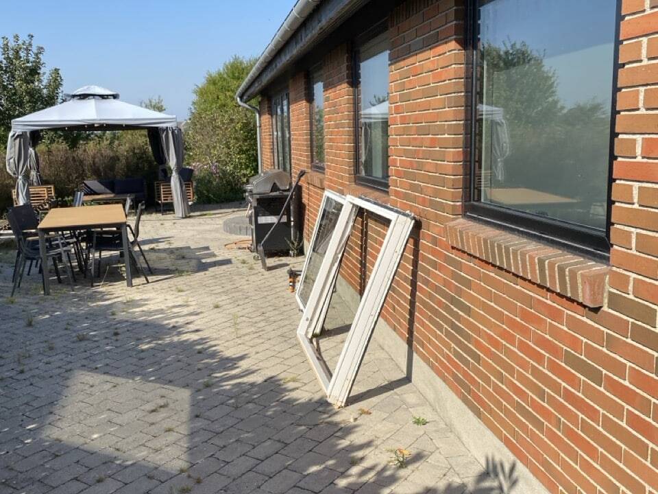 Et murstenshus med store vinduer på en solrig dag. To vinduesrammer, fremstillet af Tømrer Slagelse, læner sig op ad muren. Til venstre er der opstillet havemøbler og et lysthus på et asfalteret område omgivet af grønt.