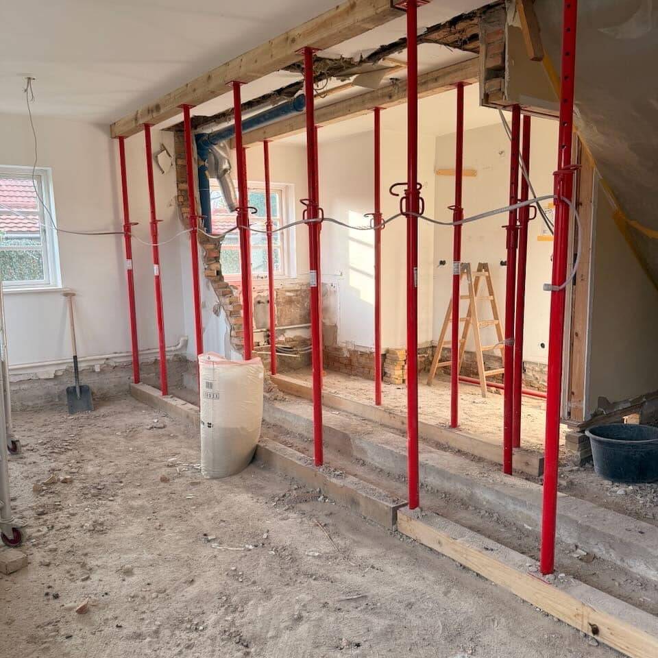 Et rum under renovering med synlige gulve og røde stålstøtter, der holder loftet oppe. Byggematerialer, værktøj fra en tømrer i Slagelse, en stige og murbrokker er synlige i hele rummet.