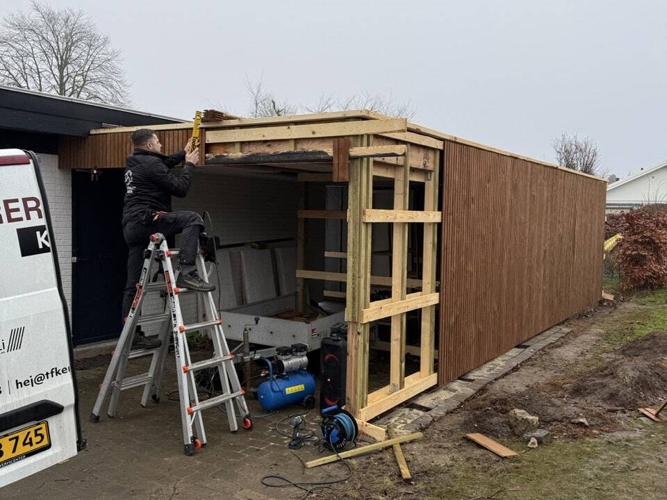 En person står på en stige og arbejder på trærammen til en carport eller et skur ved siden af et hus. Byggeredskaber og materialer ligger spredt rundt omkring, og en varevogn er parkeret i nærheden. Strukturen er delvist bygget med træpaneler.